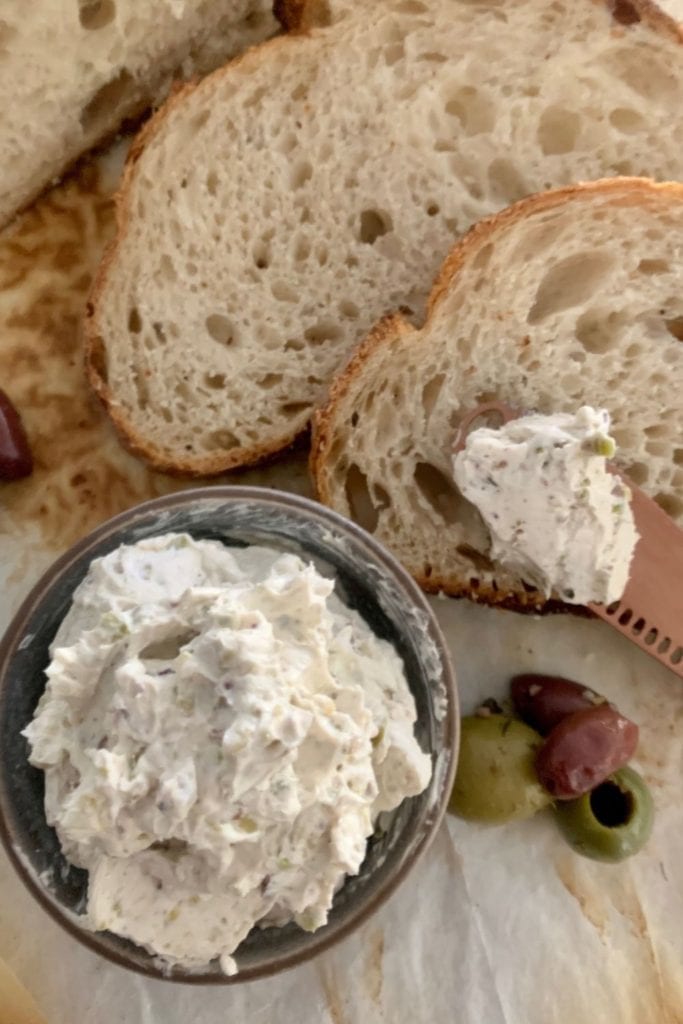 Queso crema de oliva para untar servido en un tazón pequeño.  Hay un cuchillo de mantequilla de oro rosa y pan de masa fermentada en la foto.