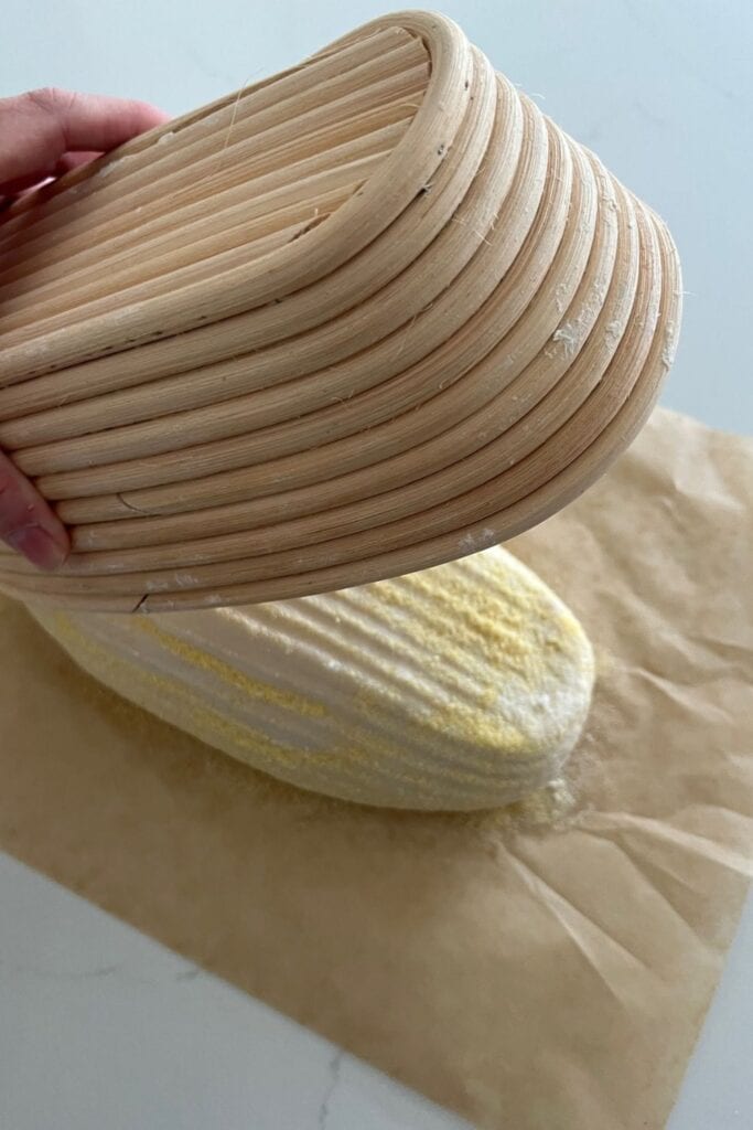Pan de masa fermentada espolvoreado con harina de maíz que se vuelca de una canasta de banneton sobre un trozo de papel pergamino.