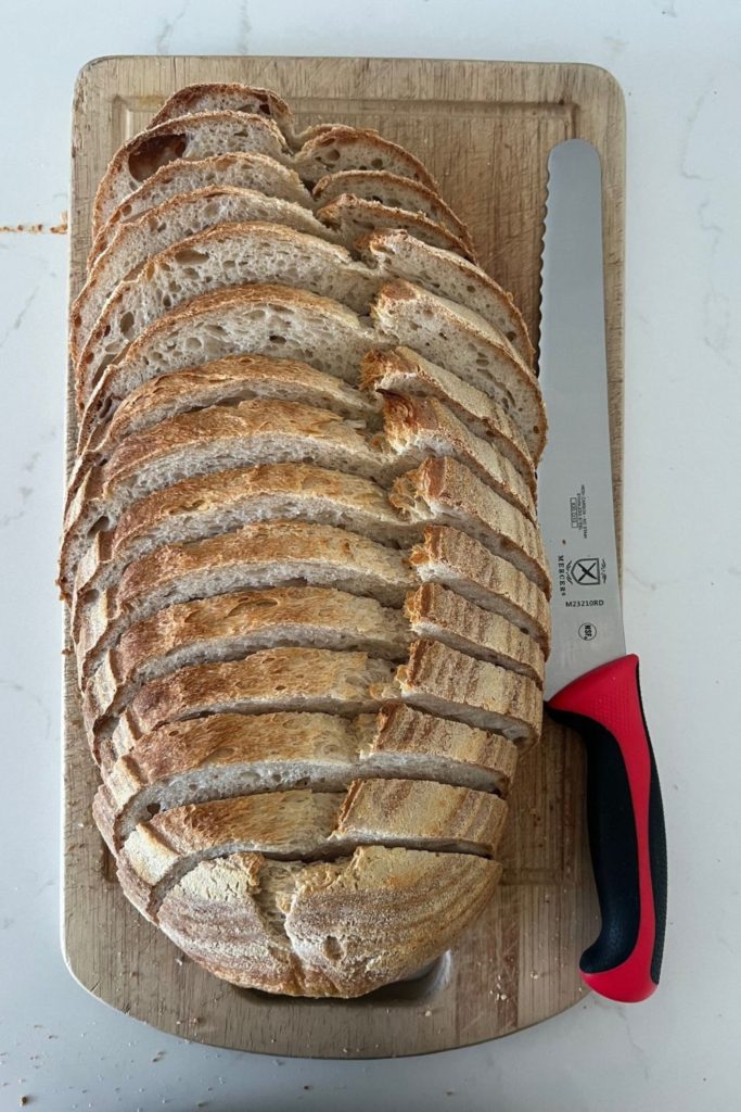 El mejor cuchillo de pan para pan de masa fermentada: cuchillo de pan Mercer con mango rojo colocado junto a una hogaza de pan de masa fermentada perfectamente rebanado.