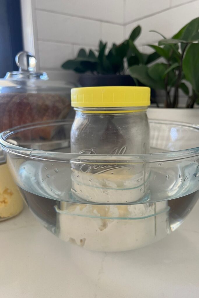 Tarro de masa fermentada con tapa amarilla en un recipiente con agua tibia.  Hay plantas verdes detrás del cuenco y una hogaza de pan de masa fermentada y un tarro de mantequilla en el fondo.