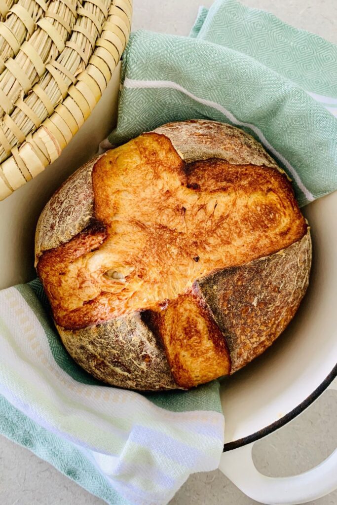 Una hogaza de pan vegano de masa fermentada dentro de un horno holandés de esmalte crema.  También hay un paño de cocina verde pálido y una cesta de caña en la foto.