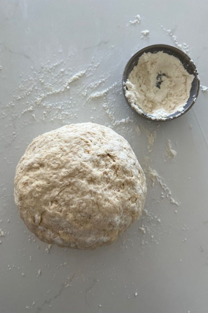 Sourdough Soda Masa de pan formada en una bola rugosa.  Hay harina en la encimera y un tazón pequeño de harina sentado a la derecha de la masa.